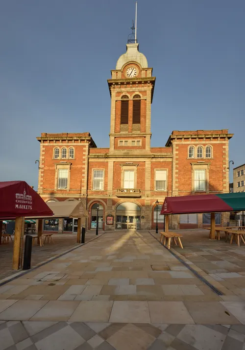 Chesterfield Market square sunrise close The image shows Chesterfield Market Square in Derbyshire, England, at sunrise during early autumn. The photograph is an urban architectural shot that prominently features the historic Chesterfield Market Hall, a well-known landmark located in the heart of Chesterfield, United Kingdom. The Market Hall is characterised by its red brick facade, clock tower, and arched windows. The scene includes market stalls with canopy coverings and picnic benches set up for the market, which is a focal point of community life in Chesterfield. The soft golden morning light casts long shadows across the newly paved square, highlighting the textures of the stone and the architectural details of the Market Hall. The image conveys the tranquil atmosphere of the market square during dawn as preparations for the day's market begin.